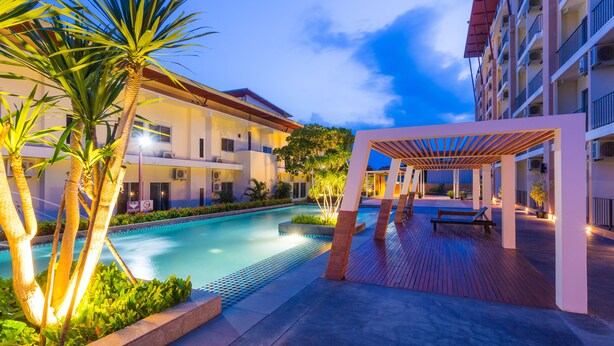A swimming pool with a wooden deck and palm trees, surrounded by a multi-story building with balconies. - Kanokan Hotel (Kanchanaburi)