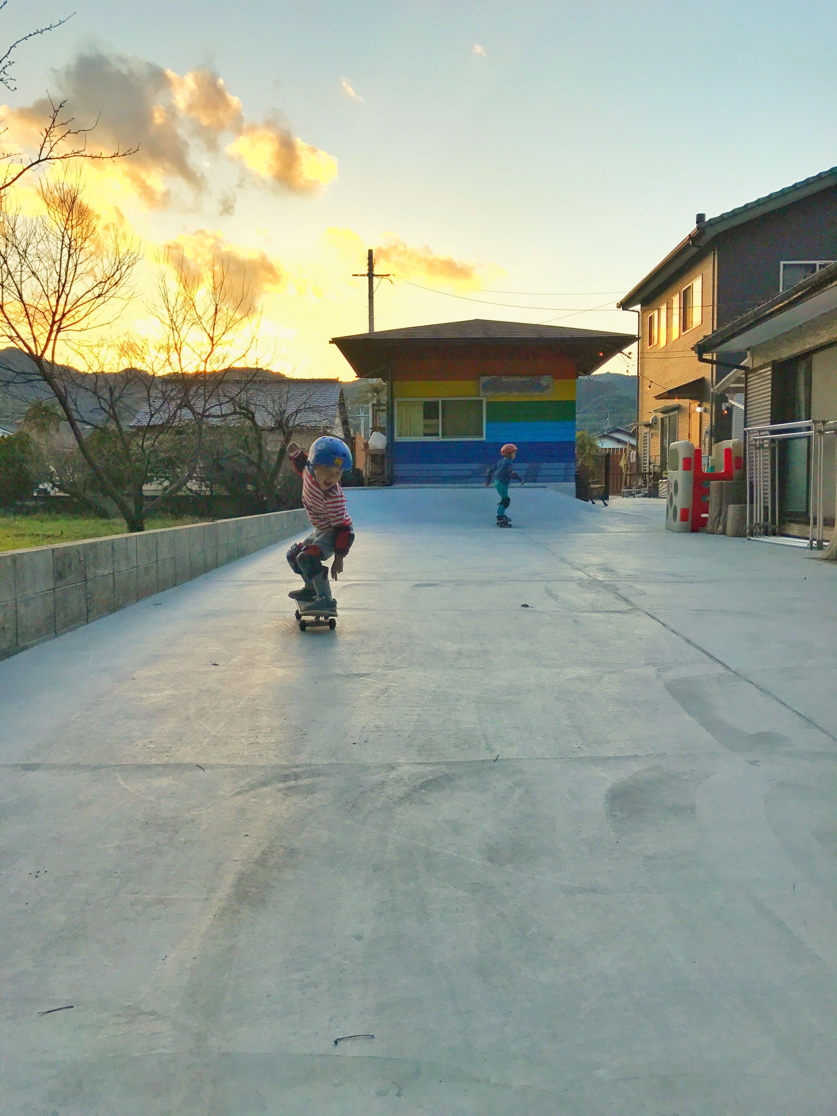 Two children skateboarding on a concrete path with buildings and a colorful structure in the background.