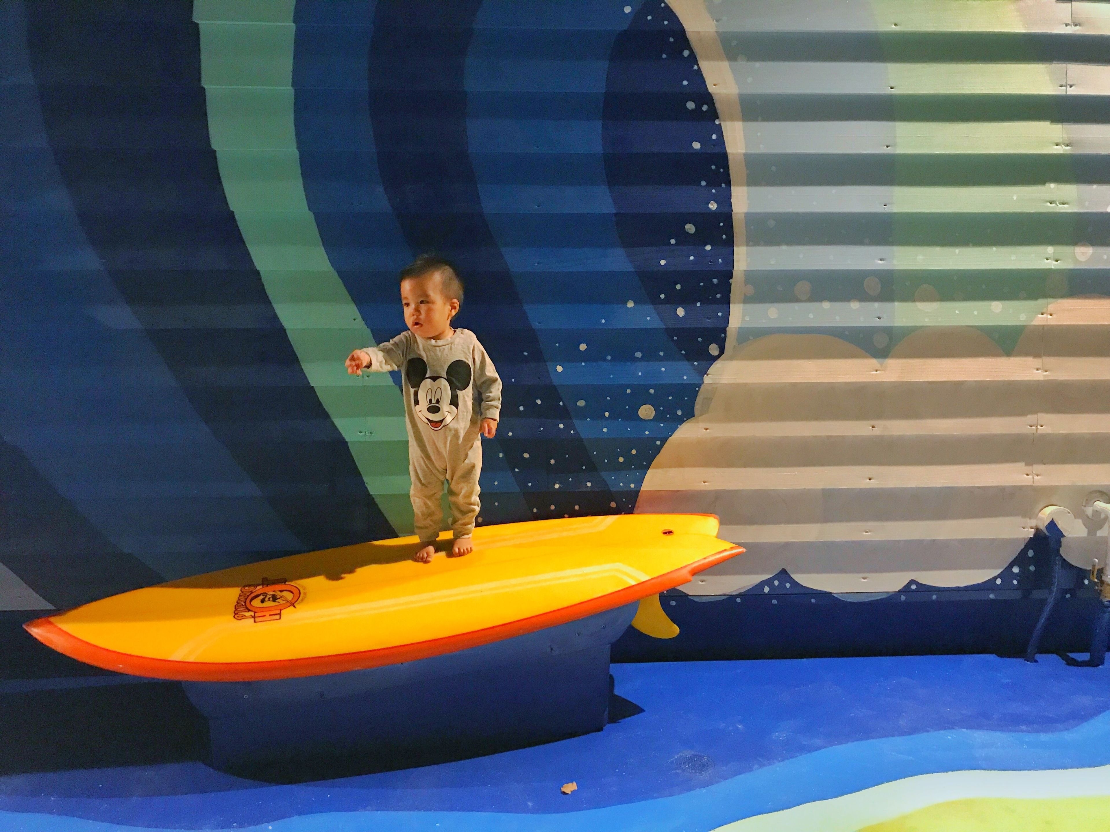 A child in a Mickey Mouse onesie standing on a yellow surfboard in a play area with a space-themed mural.