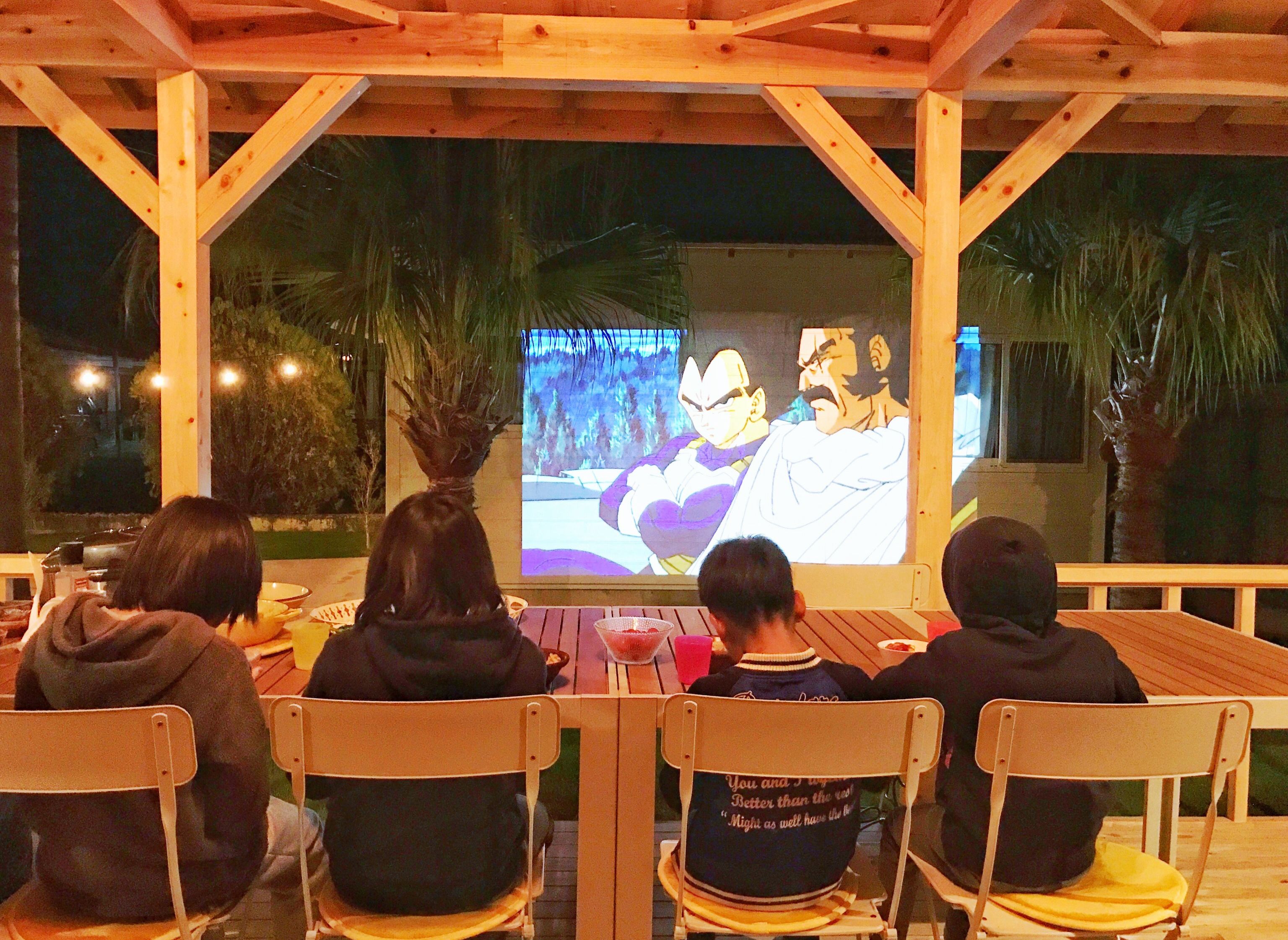 Four people seated at a wooden table under a wooden structure, watching a projected image on a screen.