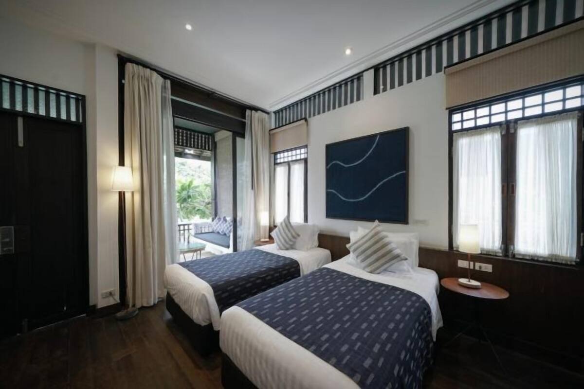 Quarto de hotel com duas camas, vista para área verde, obra de arte na parede e uma pequena mesa com luminária.