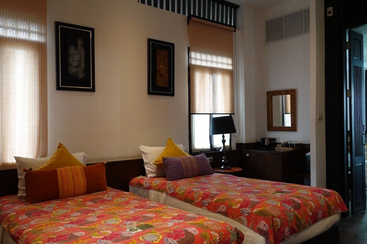 Quarto de hotel com duas camas, cada uma com roupa de cama estampada em cores vibrantes, uma mesa de cabeceira com abajur, e uma escrivaninha com espelho e um pequeno armário.