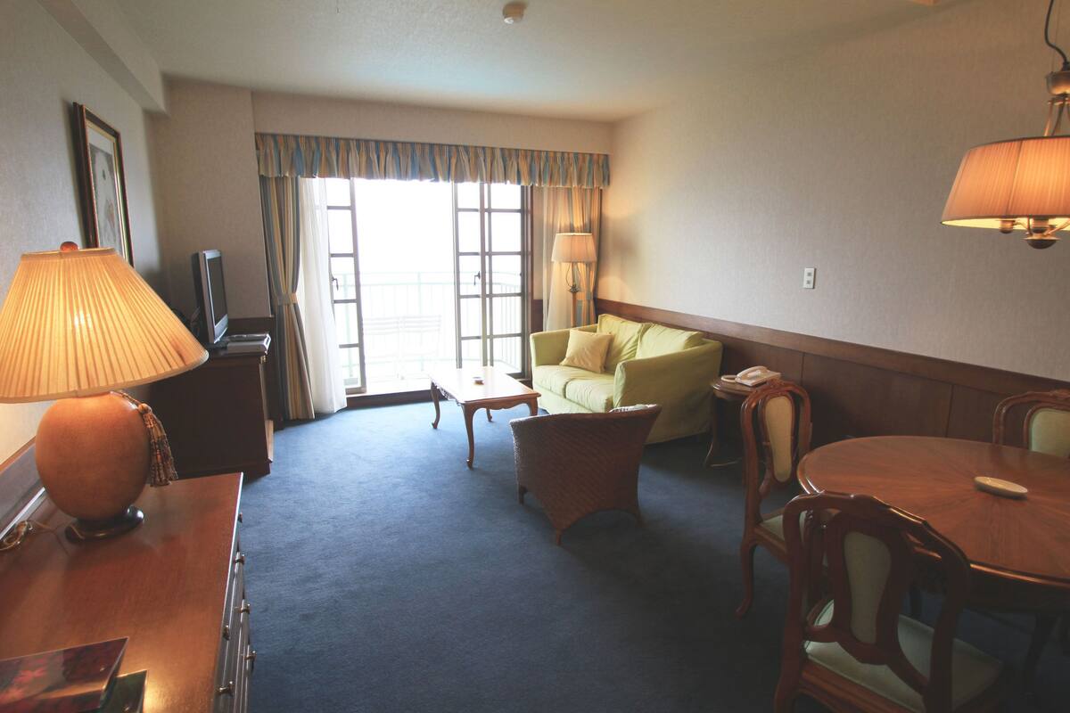 Quarto de hotel com sofá, mesa de jantar, luminária e televisão.