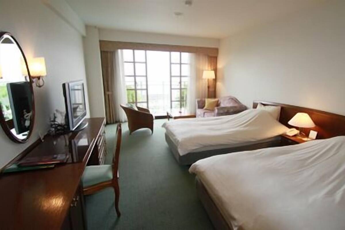 Quarto de hotel com duas camas, uma escrivaninha, uma cadeira e uma janela grande.
