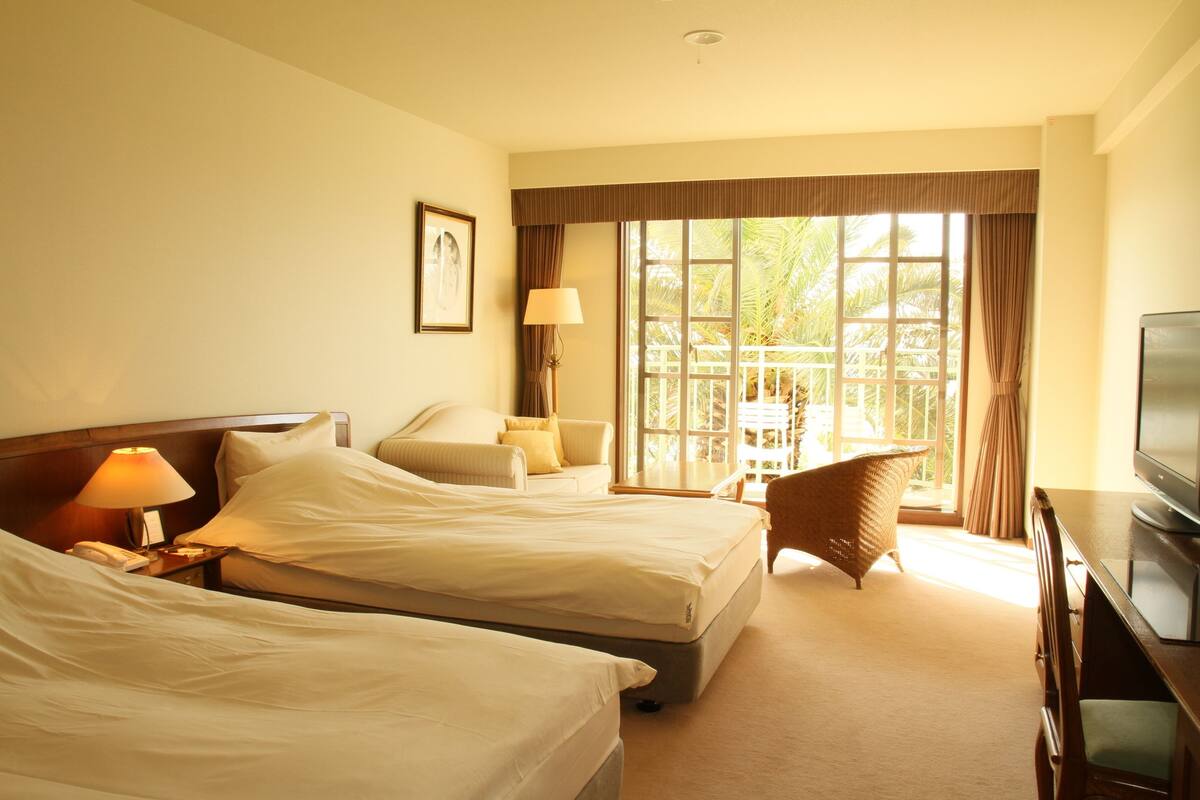 Quarto de hotel com duas camas, televisão, luminária, cadeira e janela com cortinas.