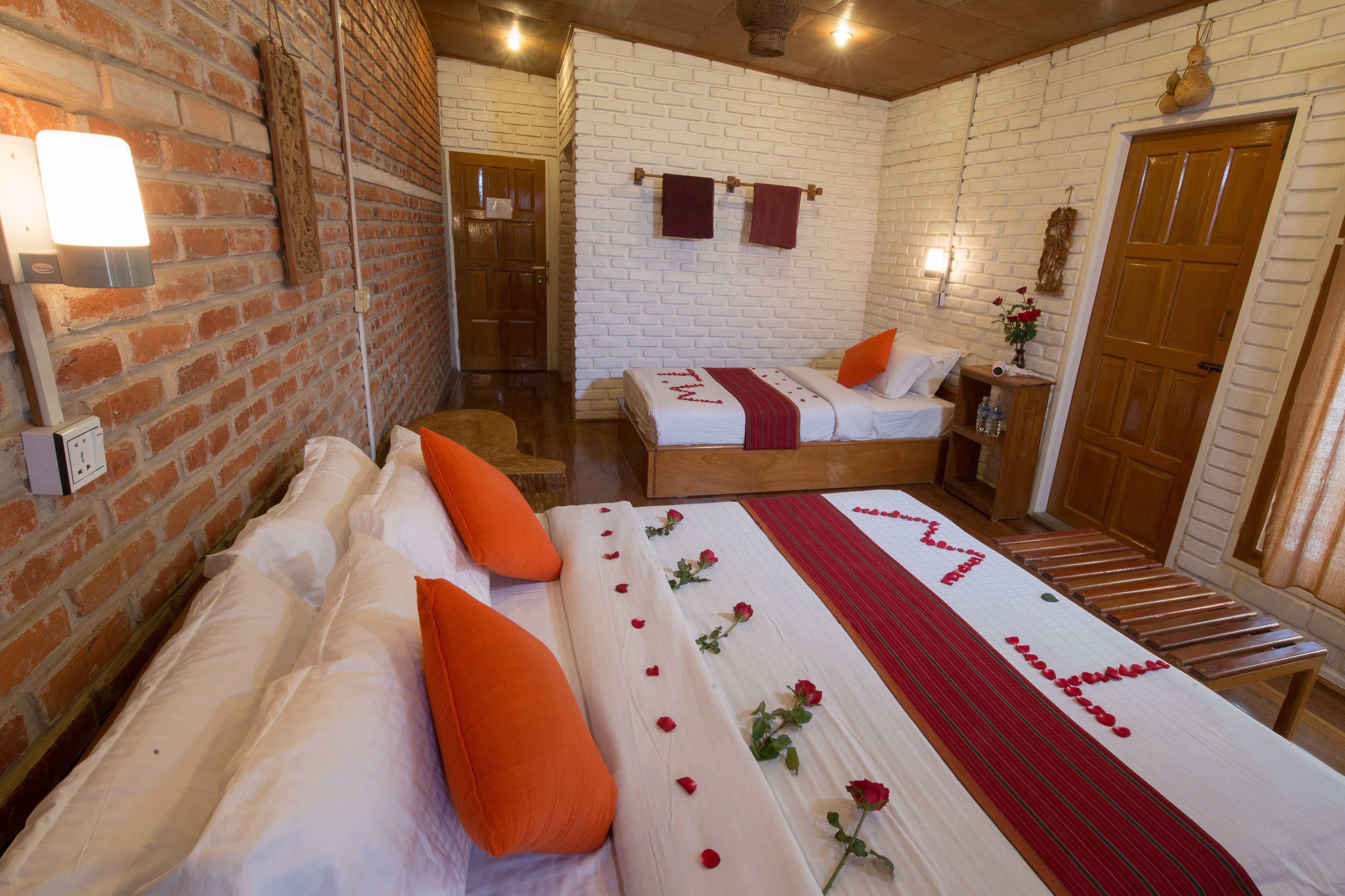 A hotel room with a brick wall, a wooden bed with orange pillows, a white bedspread with red roses, and a wooden bench.
