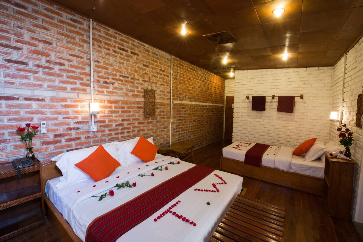 A hotel room with a brick wall, a wooden bed, a smaller bed, a bedside table with a lamp, and a mirror.