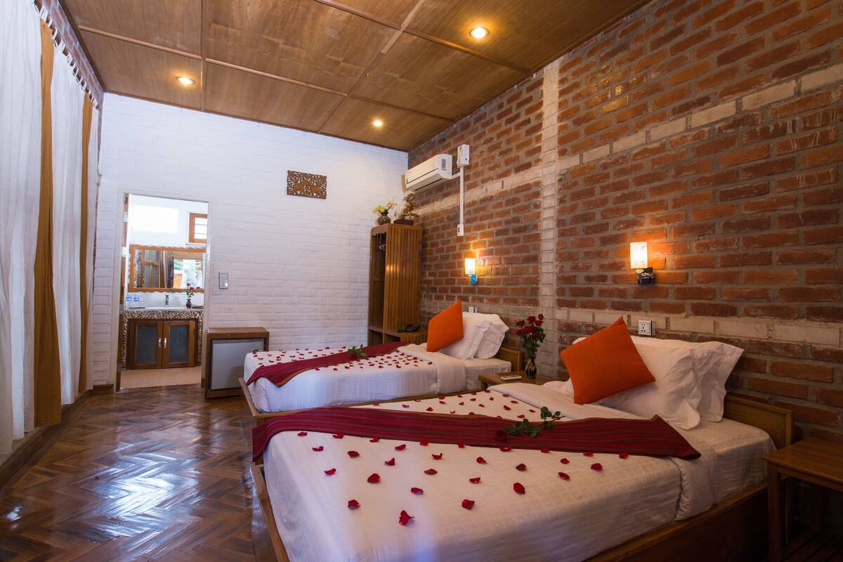 A hotel room with two beds, wooden flooring, brick walls, and a small kitchenette in the background.