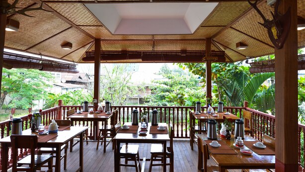 An outdoor dining area with wooden tables and chairs, surrounded by a wooden railing, and a thatched roof overhead. - Teak Wood Hotel (Nyaungshwe)