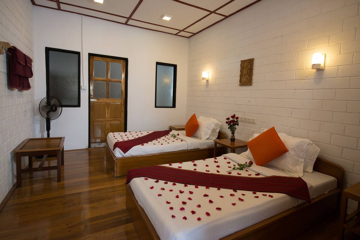 A hotel room with two beds, wooden flooring, a fan, a wooden bench, and a door.