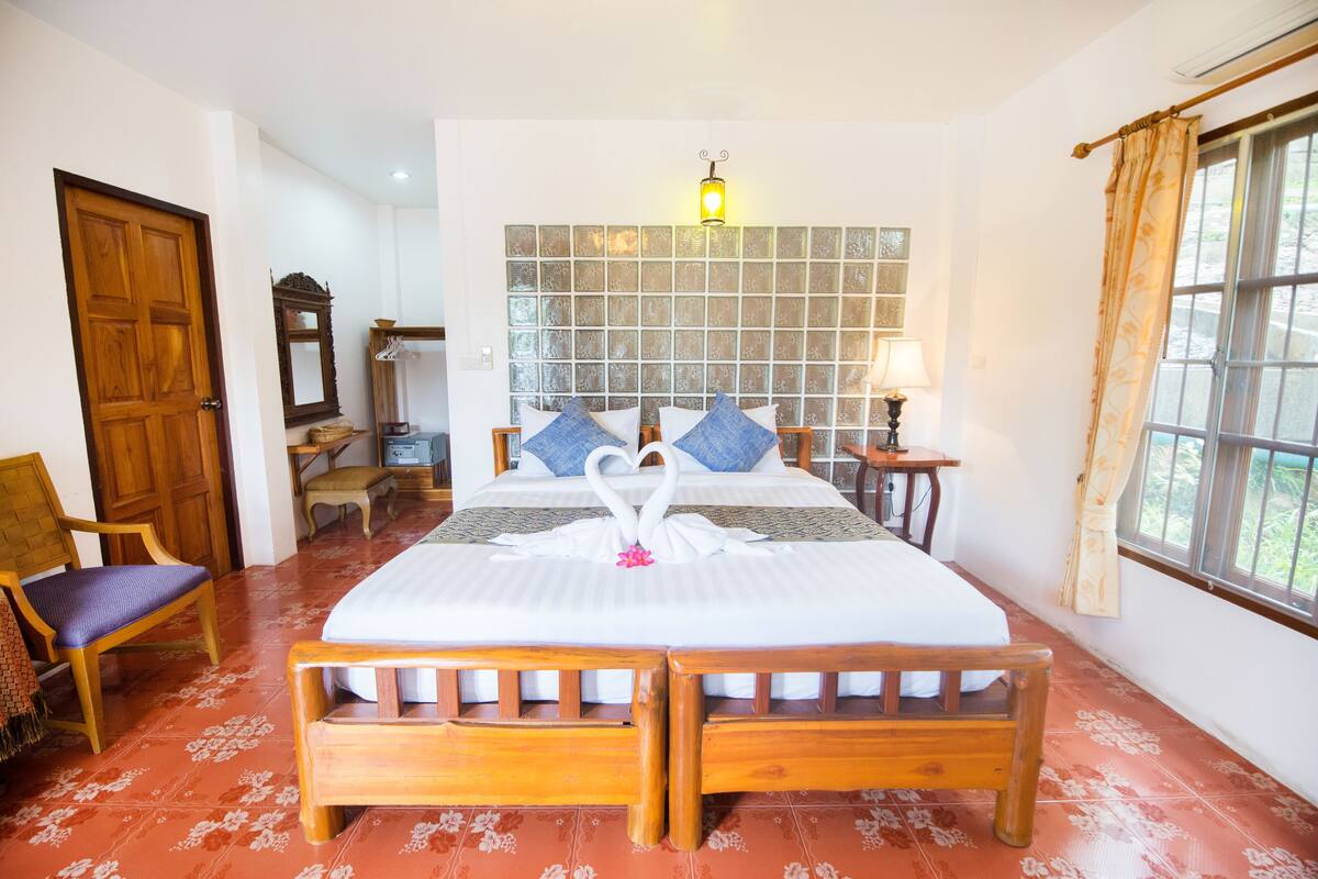 Quarto de hotel com uma cama grande, cabeceira de madeira, espelho, cadeira, mesa de cabeceira e janela com cortinas.