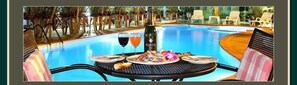 Un área de comedor junto a la piscina con una mesa para dos, una botella de champán y una copa de vino tinto.
