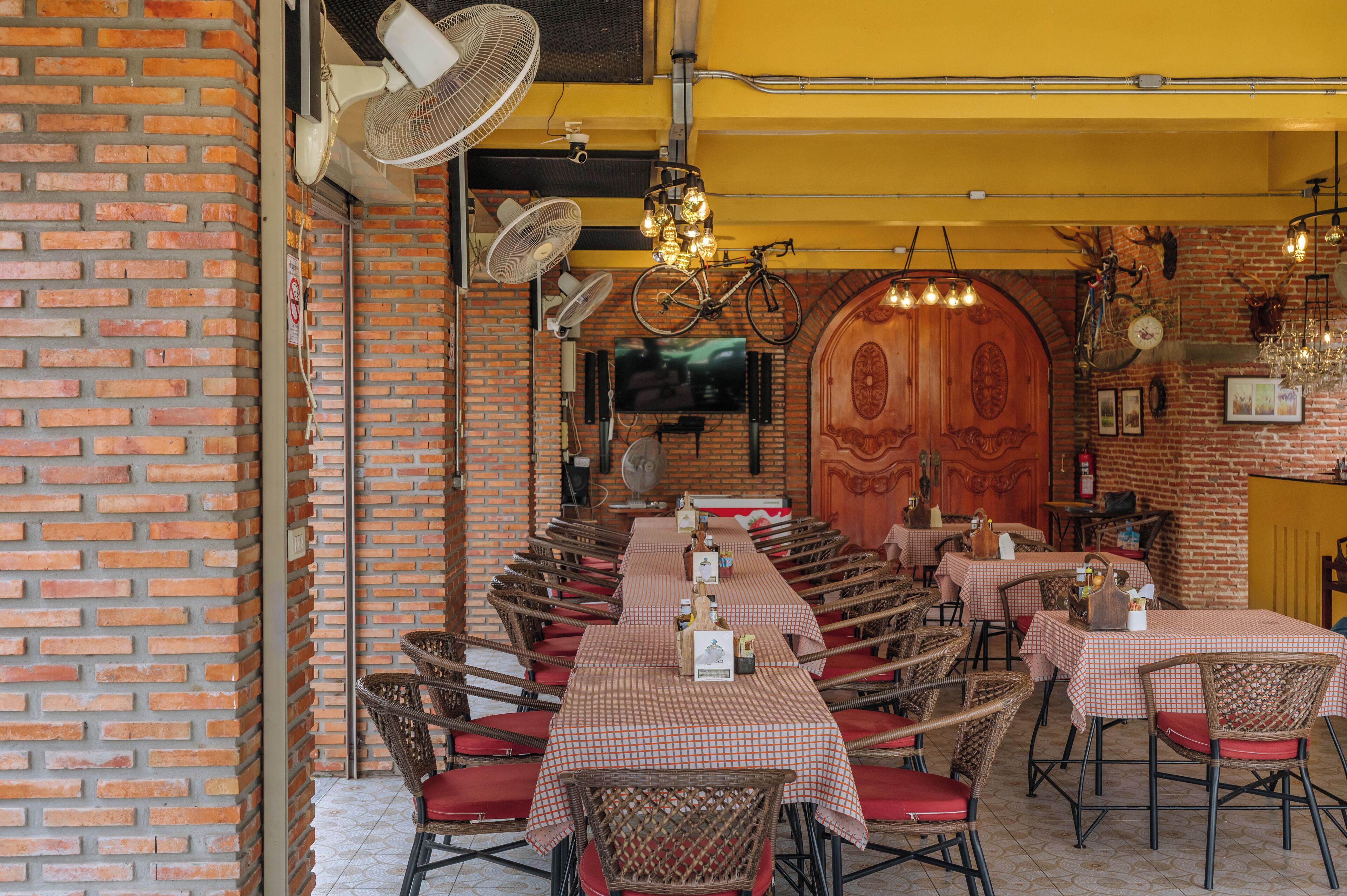 A restaurant with brick walls, multiple tables with checkered tablecloths, wicker chairs, and a large wooden door.