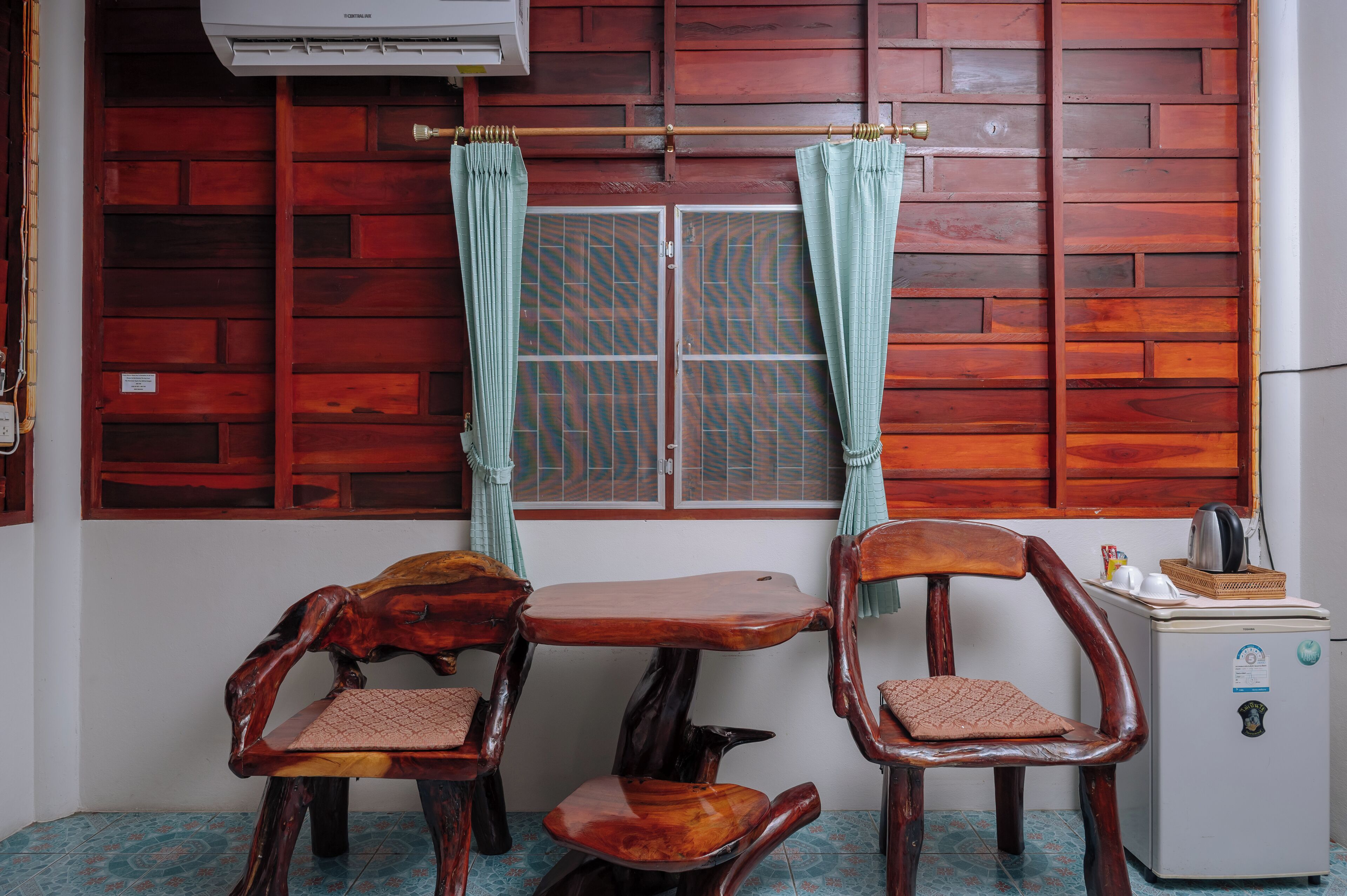 Una habitación con pared de madera, una ventana con cortinas, una mesa pequeña, dos sillas y un refrigerador.