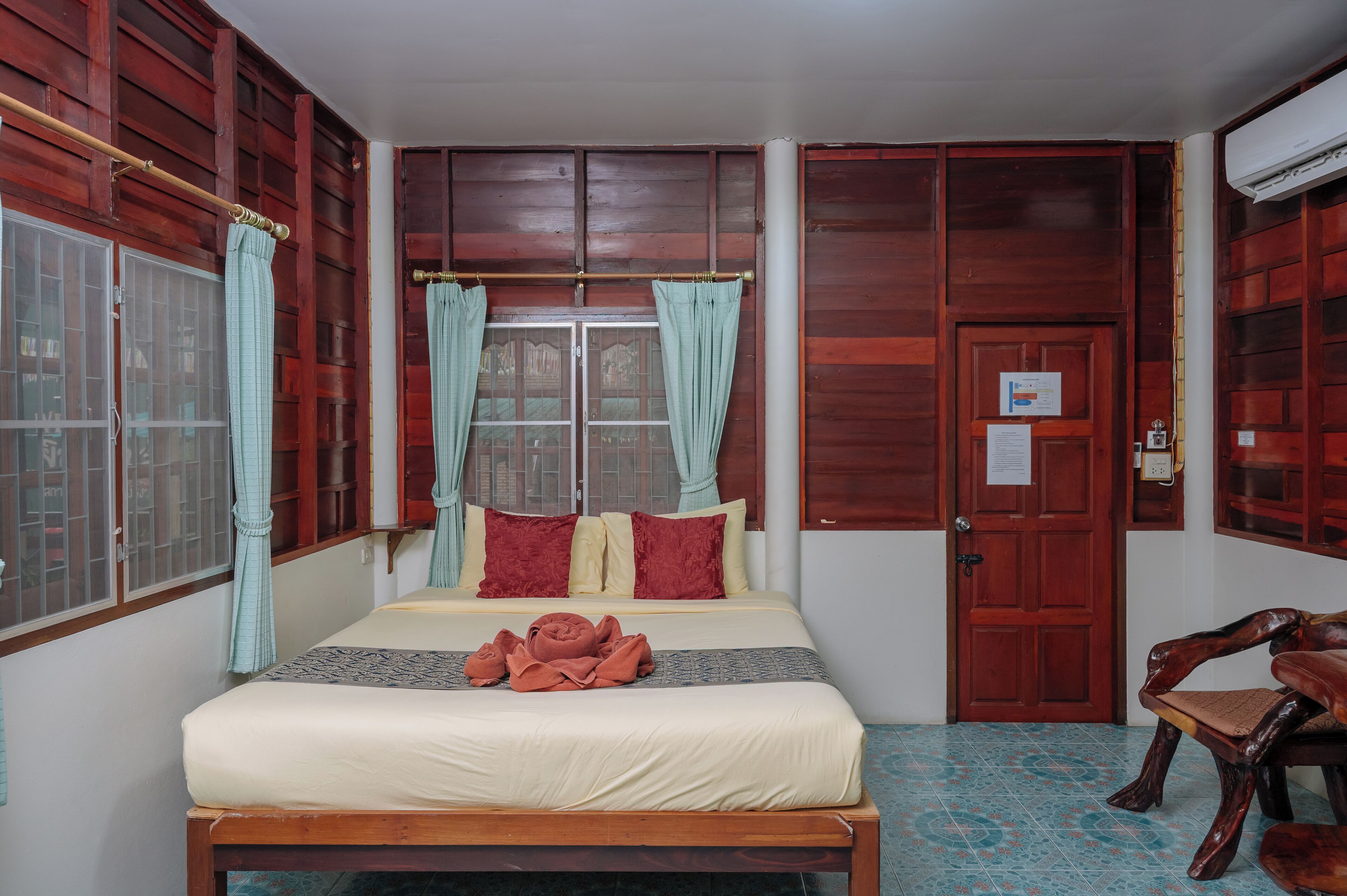 A room with a bed, a wooden bench, a window with curtains, and a door with a notice on it.