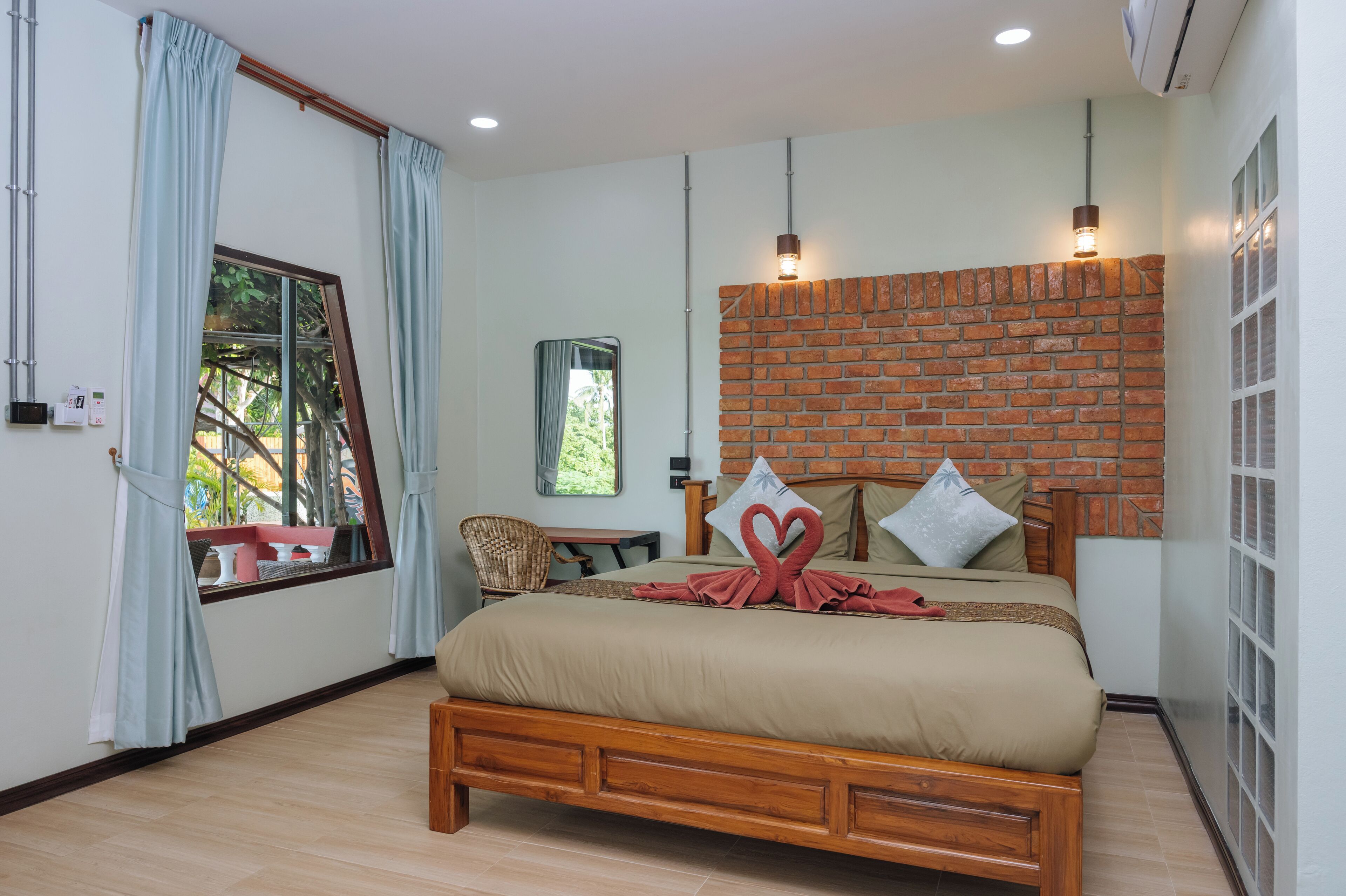 A bedroom with a brick wall, a bed with a red heart decoration, a window with curtains, and a small table with a chair.