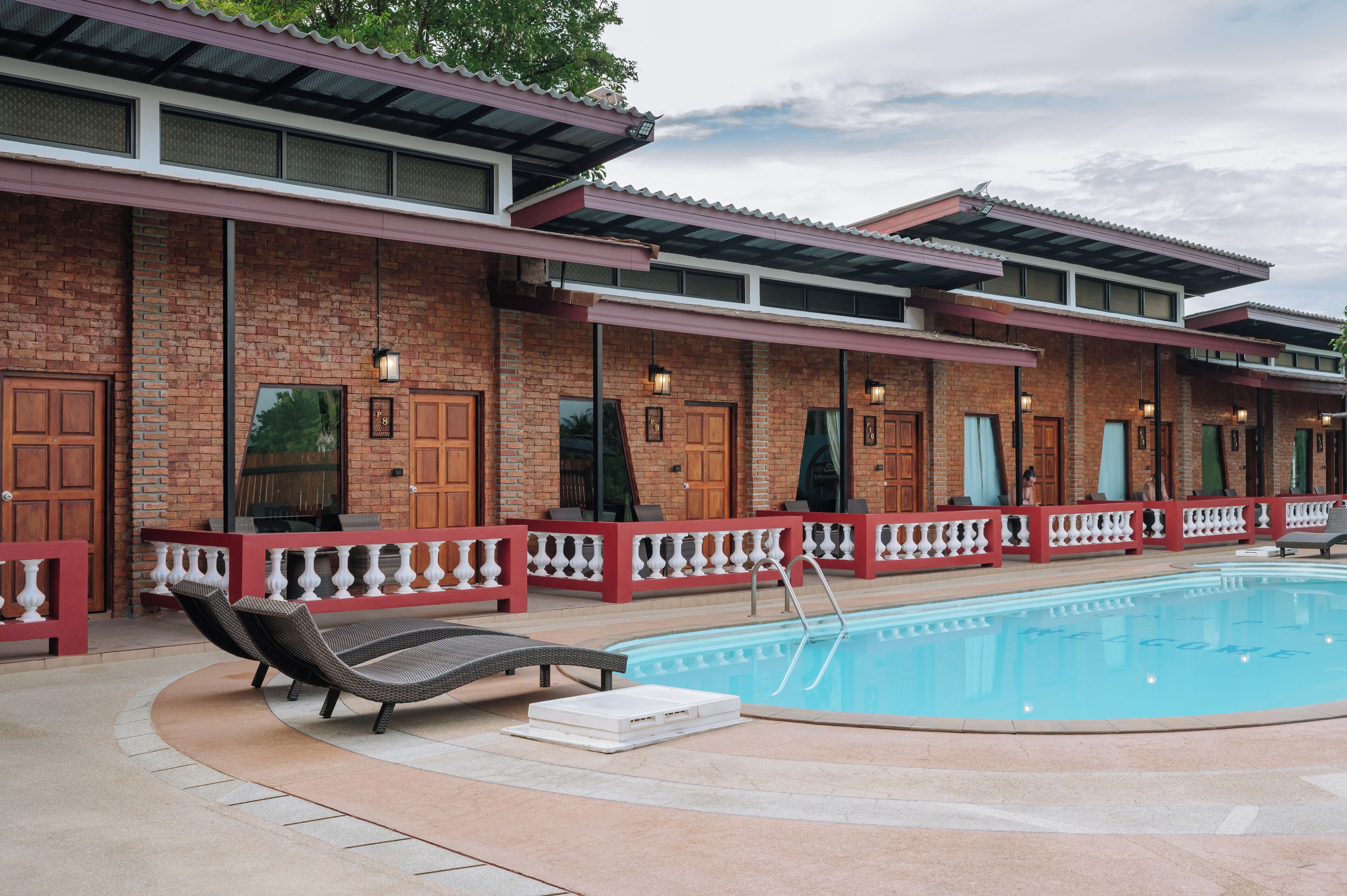 A poolside area with lounge chairs, a pool, and a row of brick buildings with large windows and doors.