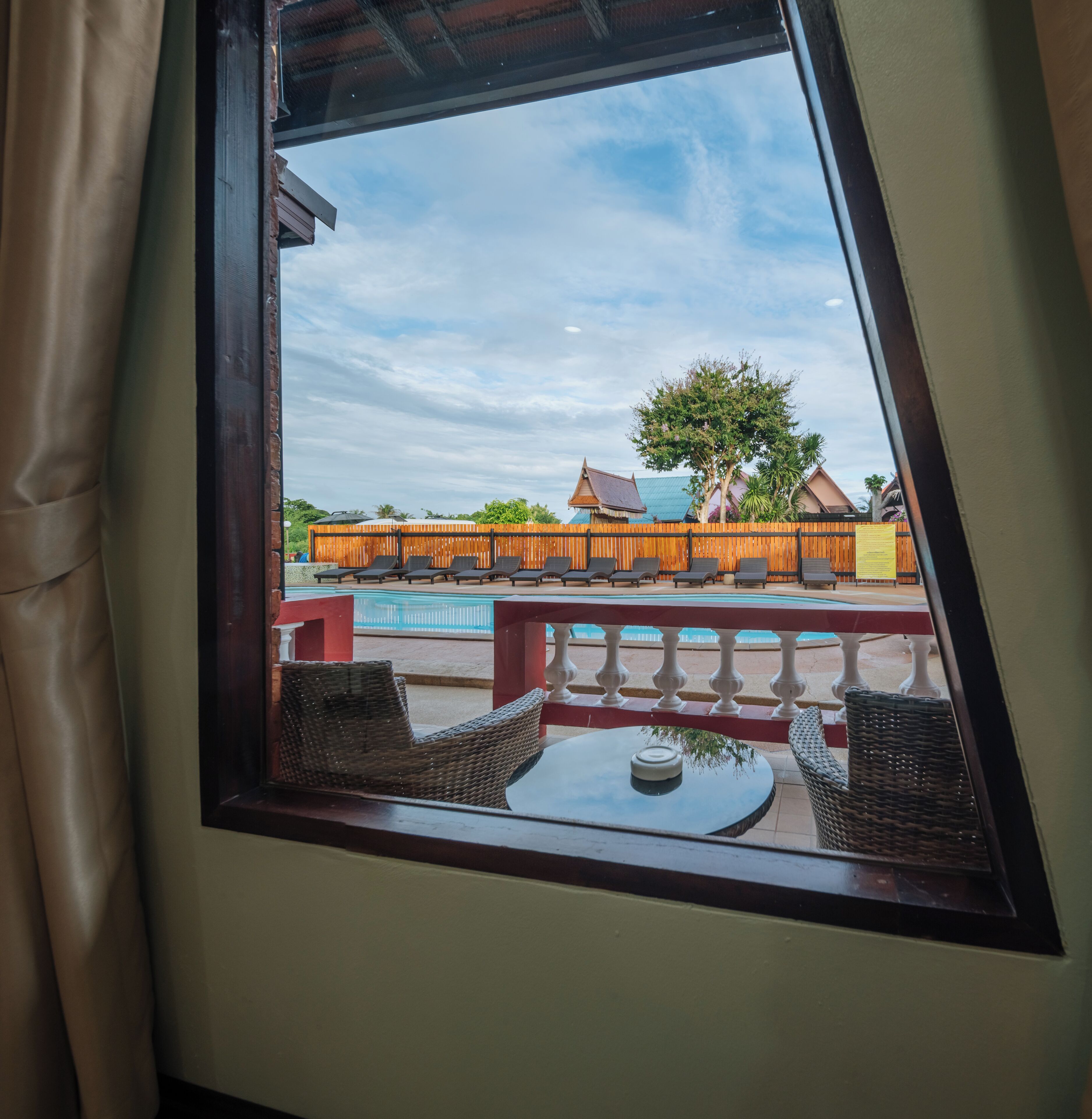 Une vue depuis une fenêtre à cadre en bois, donnant sur un espace piscine avec des chaises longues et, au loin, un bâtiment doté d’un toit végétalisé.