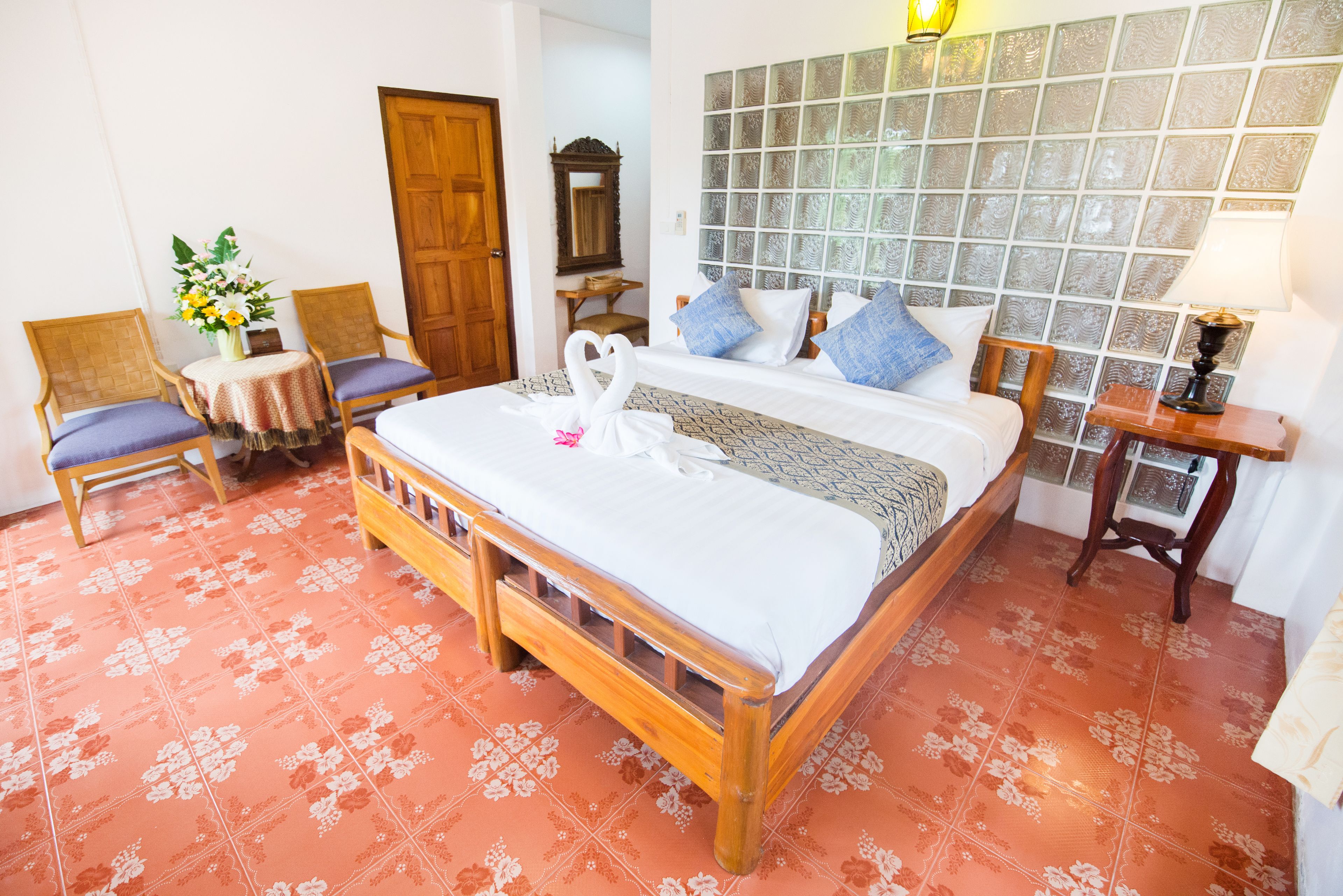 Un dormitorio ordenado con una cama de madera, dos sillas, una mesita con un jarrón de flores y un espejo en la pared.