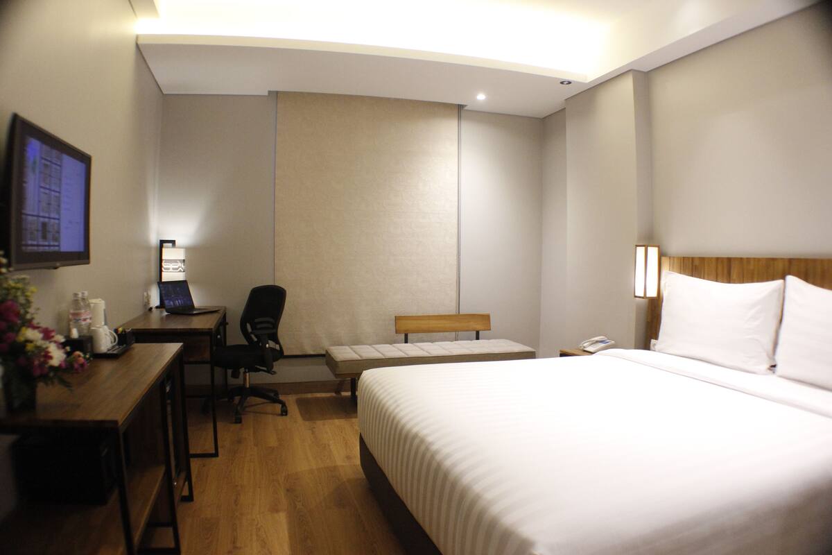 Quarto de hotel com cama, escrivaninha, cadeira, mesinha, televisão e abajur.