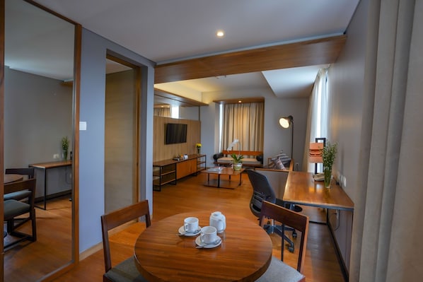 Quarto de hotel moderno com mesa de jantar de madeira para dois, um sofá, uma TV e uma cozinha.