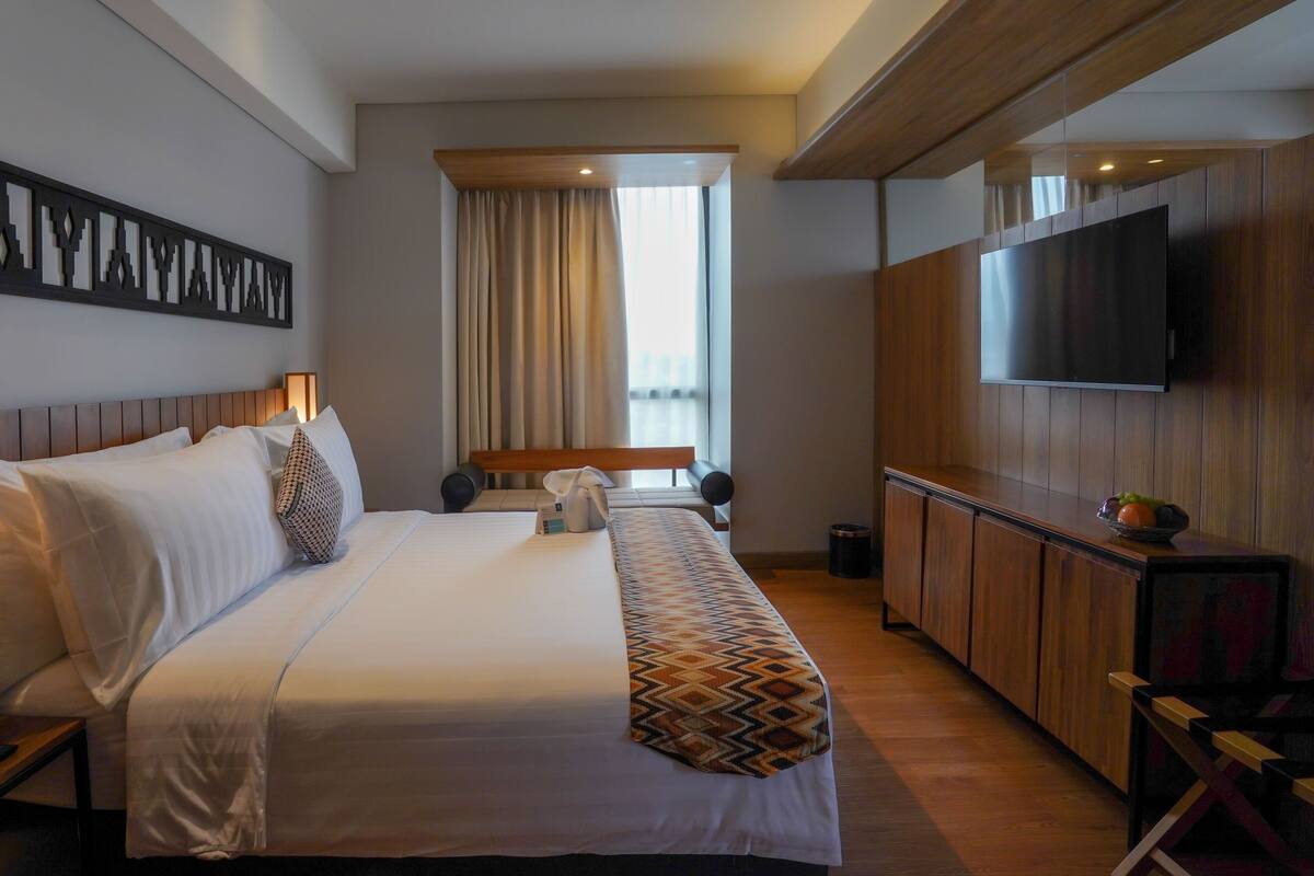 Quarto de hotel com uma cama grande, televisão montada na parede, um banco de madeira e uma janela com cortinas.