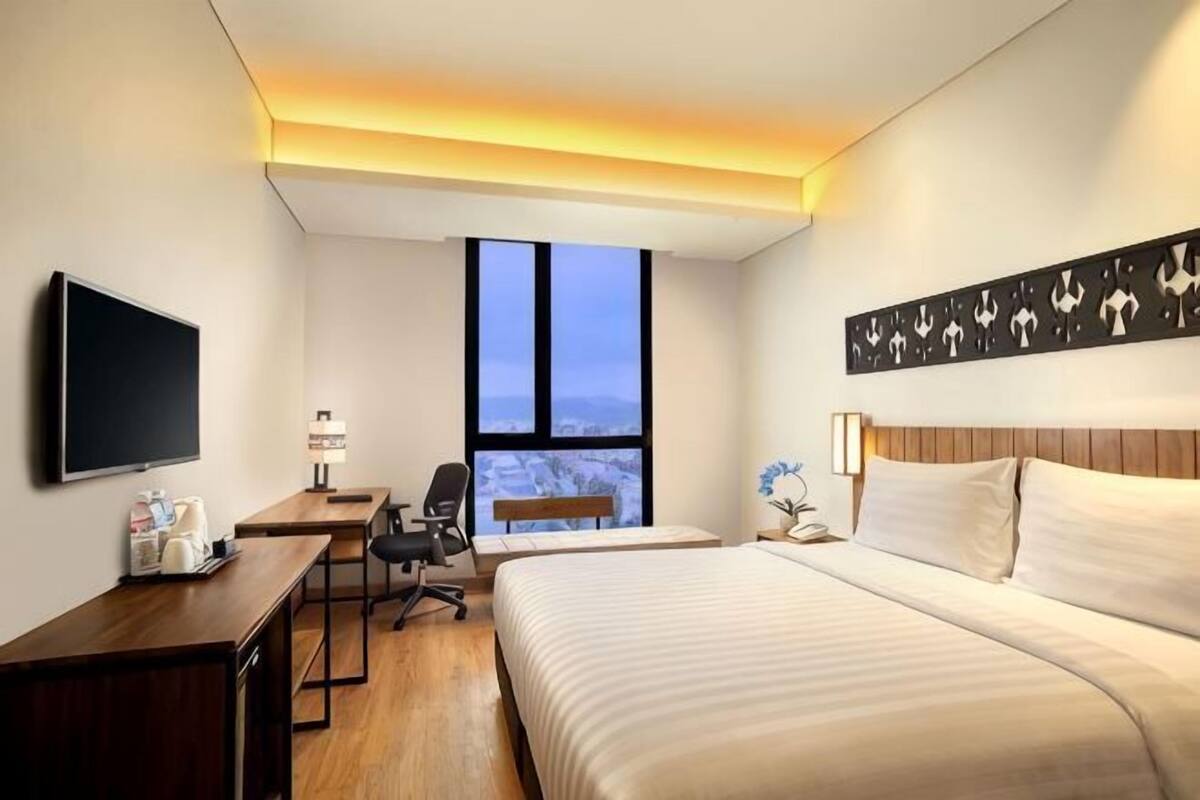 Quarto de hotel com cama, escrivaninha, cadeira, televisão e uma janela grande.