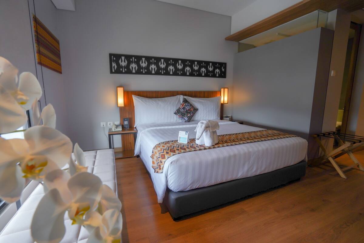 Quarto de hotel com cama grande, duas luminárias de cabeceira, travesseiro decorativo e uma obra de arte na parede.