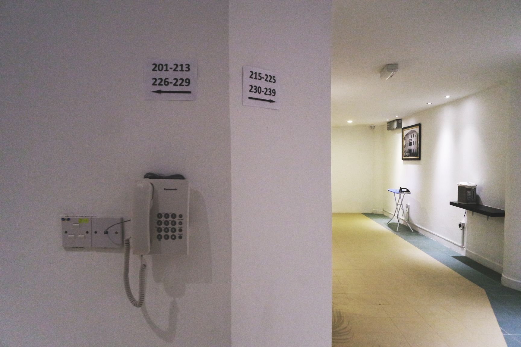 Un pasillo con letreros que indican los números de habitación desde 201 hasta 213, del 226 al 229, del 215 al 225, y del 230 al 239. Un teléfono está instalado en la pared.