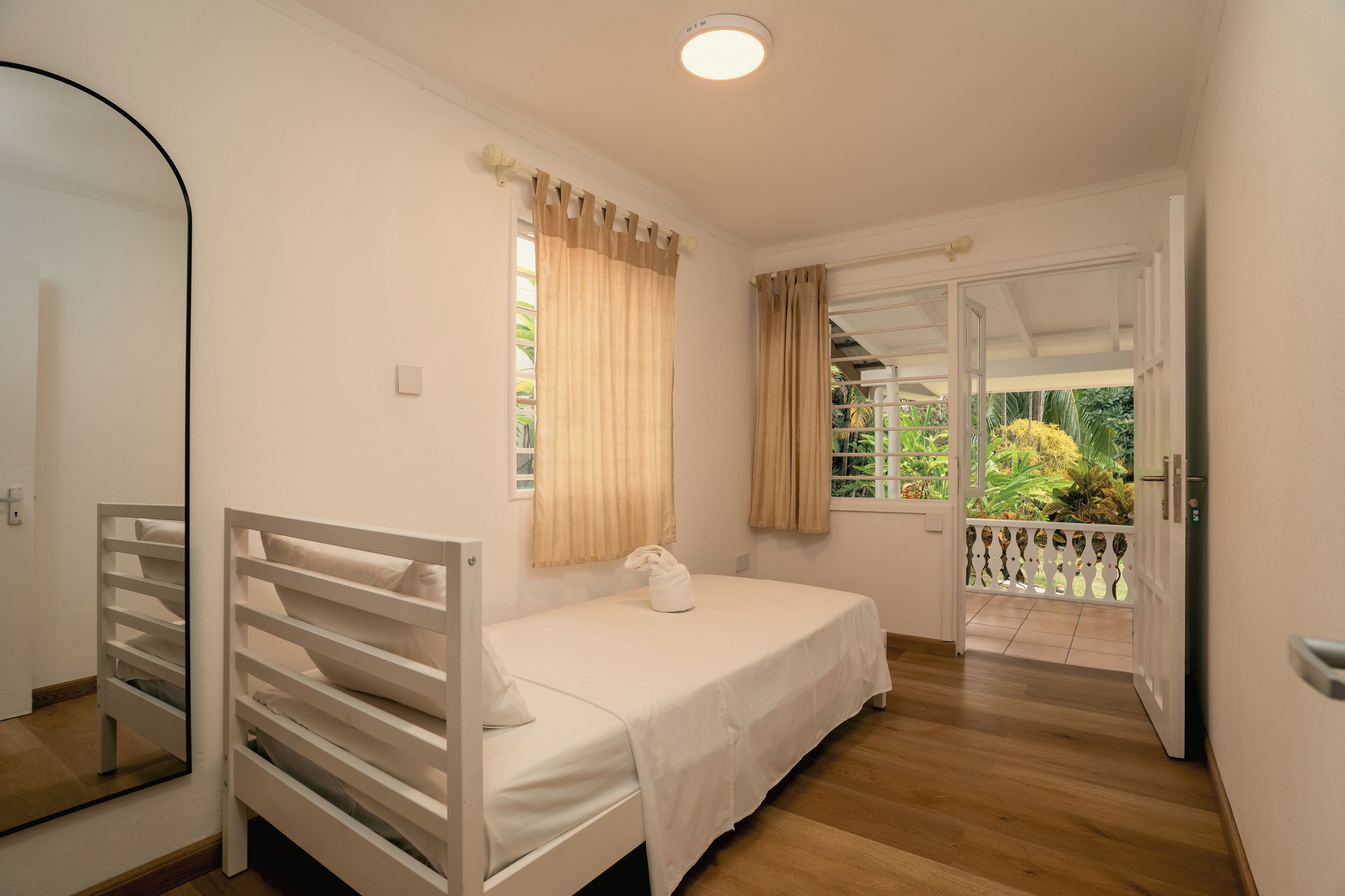 A bedroom with a single bed, a window with curtains, a door leading to a balcony, and a mirror.
