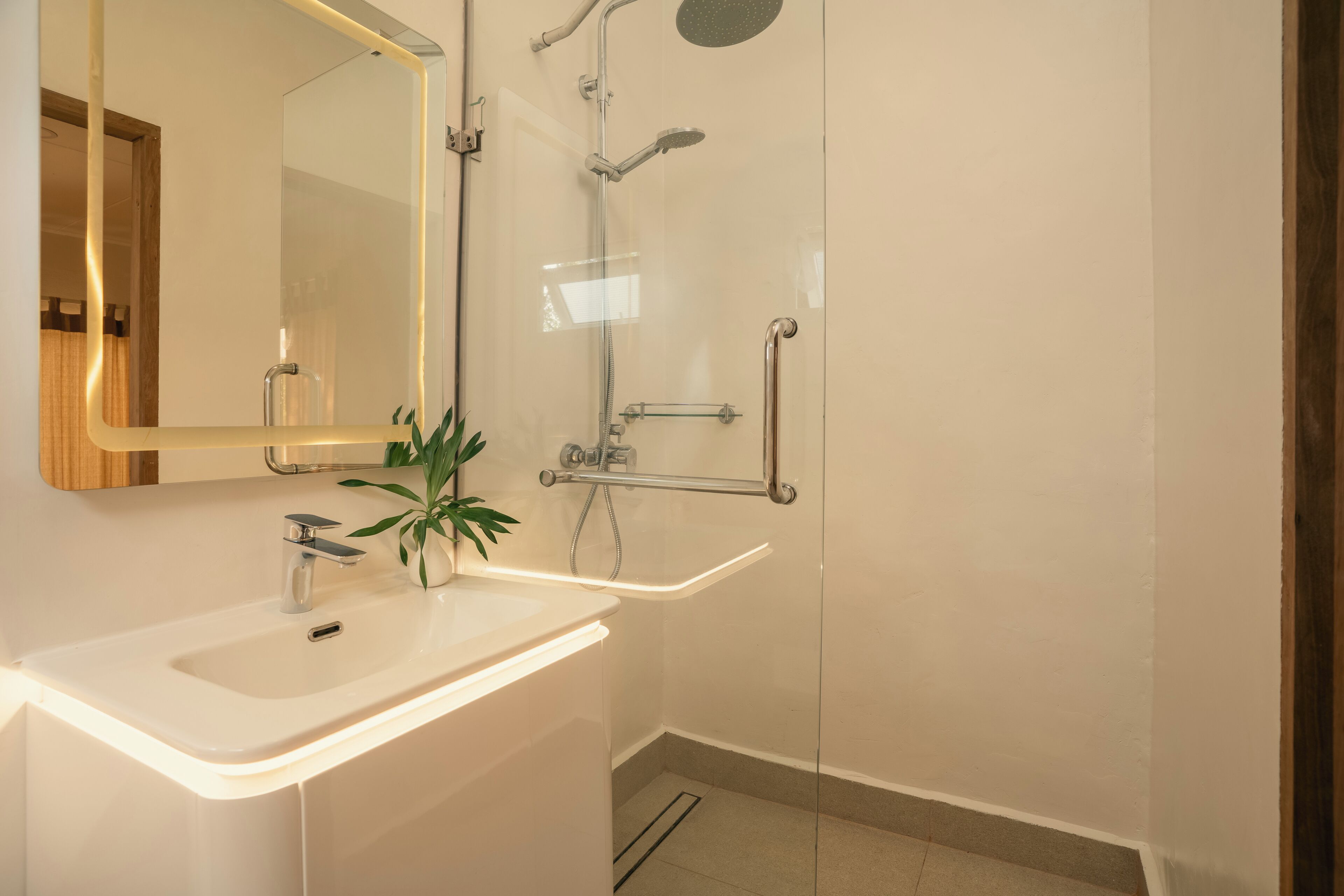 Un baño moderno con un lavabo blanco, un espejo, ducha con puerta de cristal y una planta sobre el lavabo.
