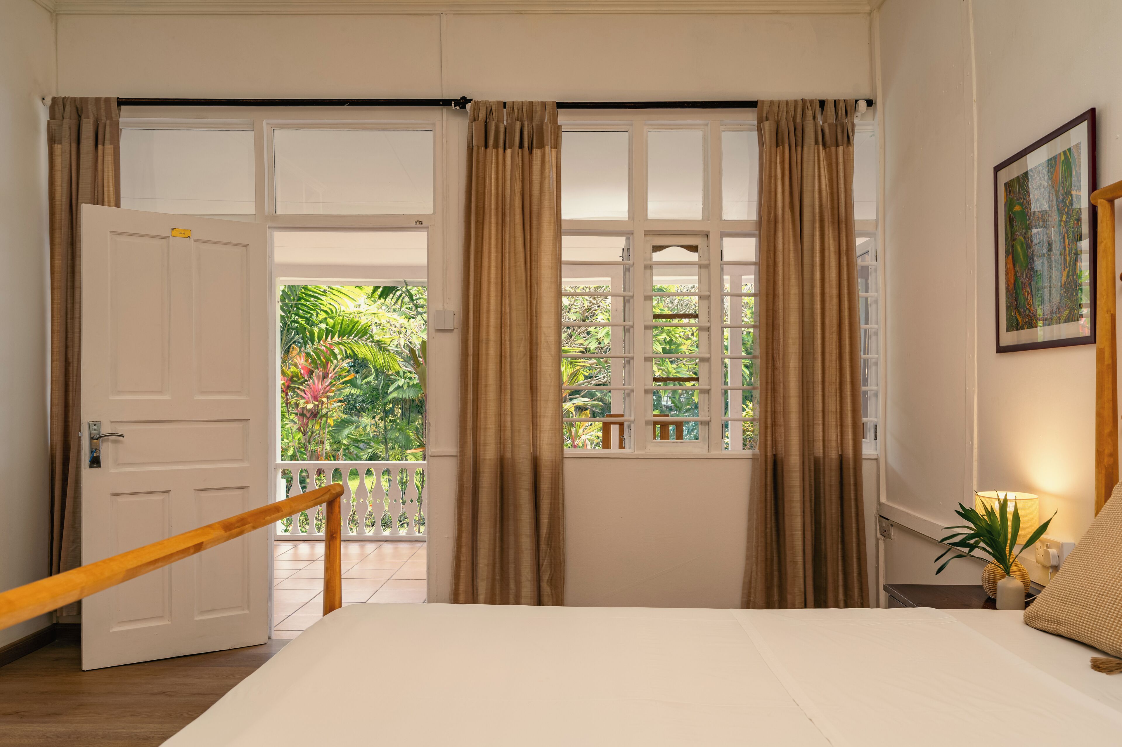 A bedroom with a large bed, a wooden railing, a balcony with curtains, and a view of greenery.