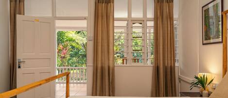 A bedroom with a large bed, a wooden railing, a balcony with curtains, and a view of greenery.