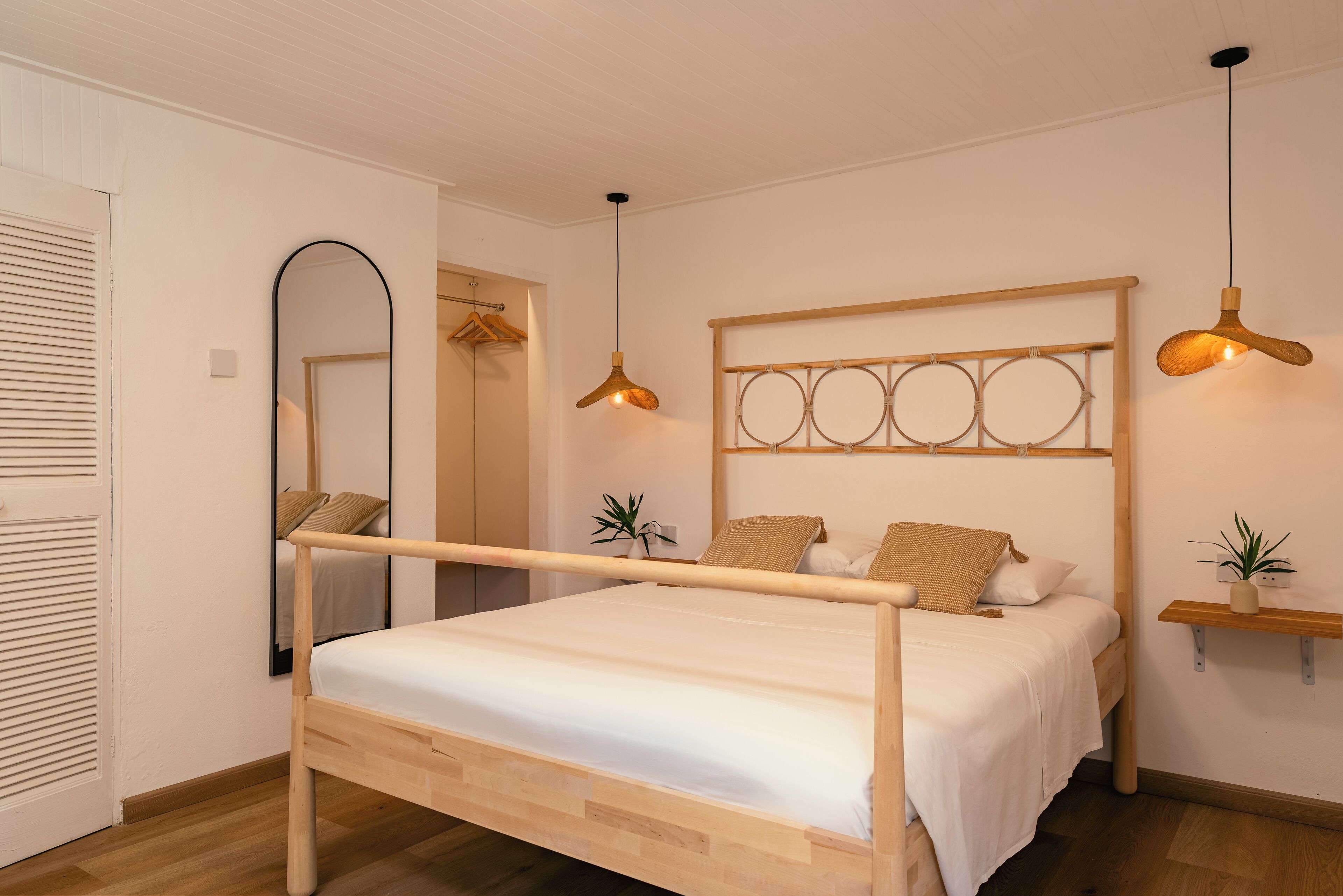 A bedroom with a wooden bed, a mirror, a hanging light fixture, and a wall-mounted shelf with a plant.