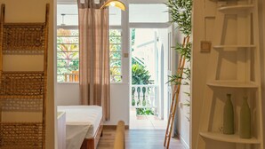 A bedroom with a bed, a bedside table, a hanging light, a window with curtains, a plant, and a white shelving unit. - Beau Vallon Bungalows (Mahé Island)