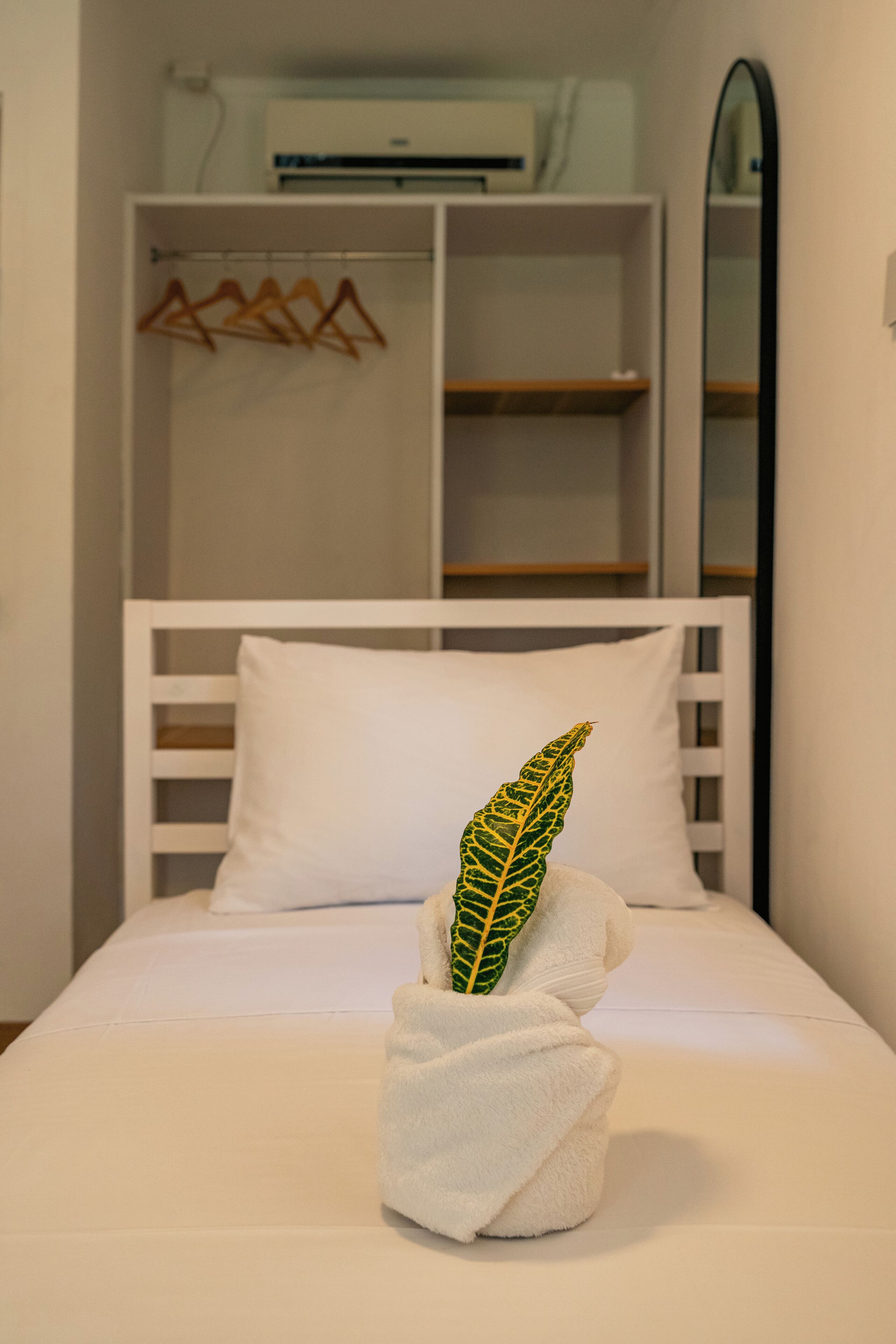 Een netjes opgemaakt bed met een wit kussen en een handdoek met een groen blad. Achtergrond is een witte kledingkast met een spiegel.