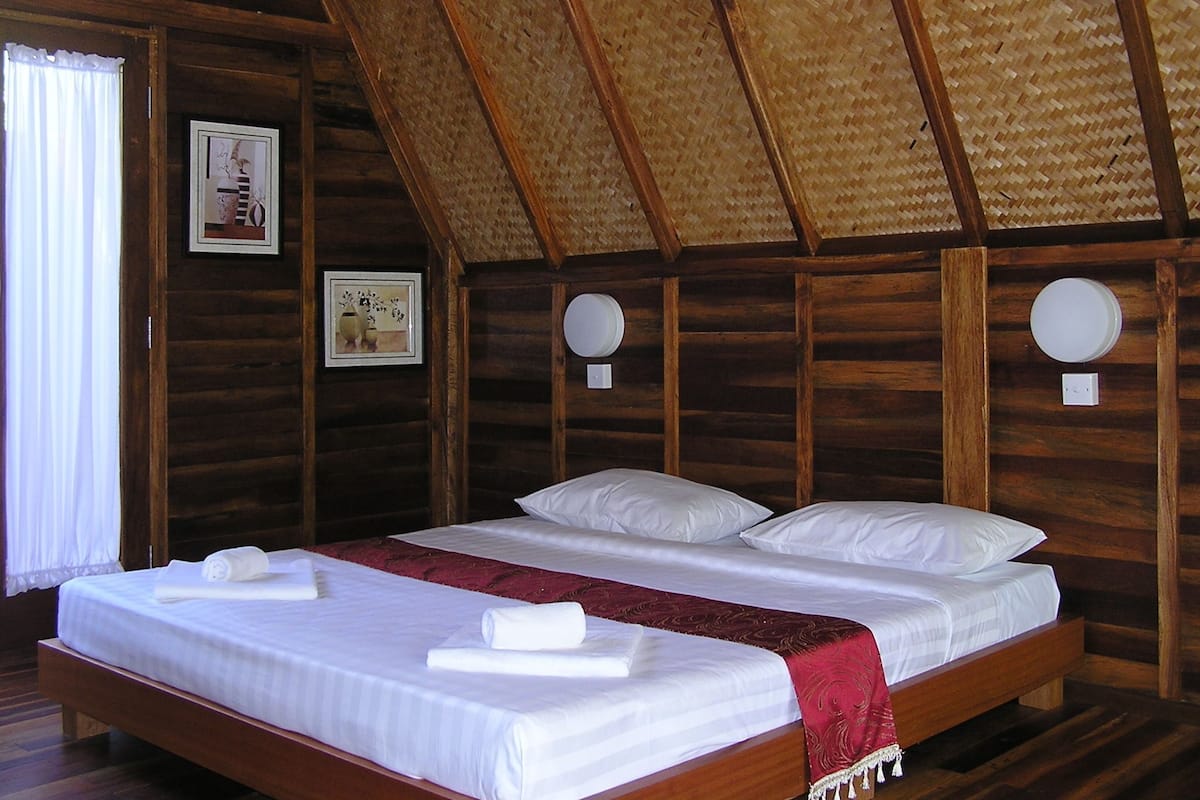 A wooden room with a high ceiling, two beds with white linens, a red blanket, and two wall-mounted light fixtures.