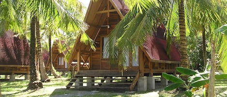 A wooden cabin with a thatched roof, surrounded by palm trees and a grassy area.