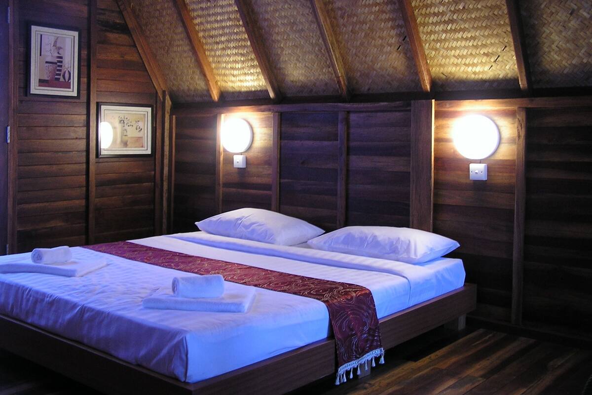A double bed with white linens and a red throw blanket, two bedside lamps, wooden walls, and a sloped ceiling.