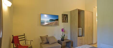 Un dormitorio con cama, una silla, una mesita con planta, armario y televisor en la pared.