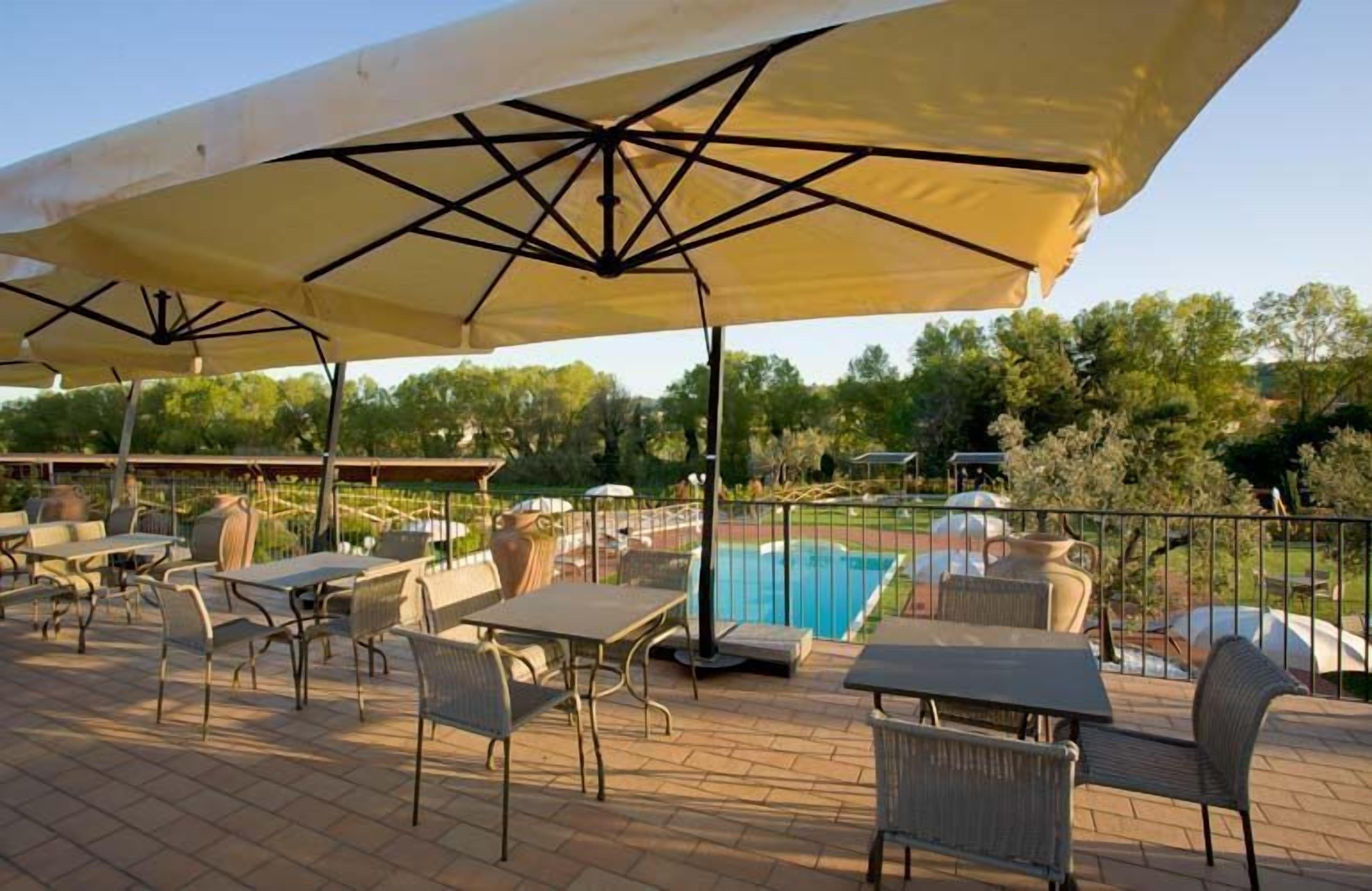Un patio al aire libre con mesas y sillas bajo un paraguas grande, una piscina y un jardín cercado.