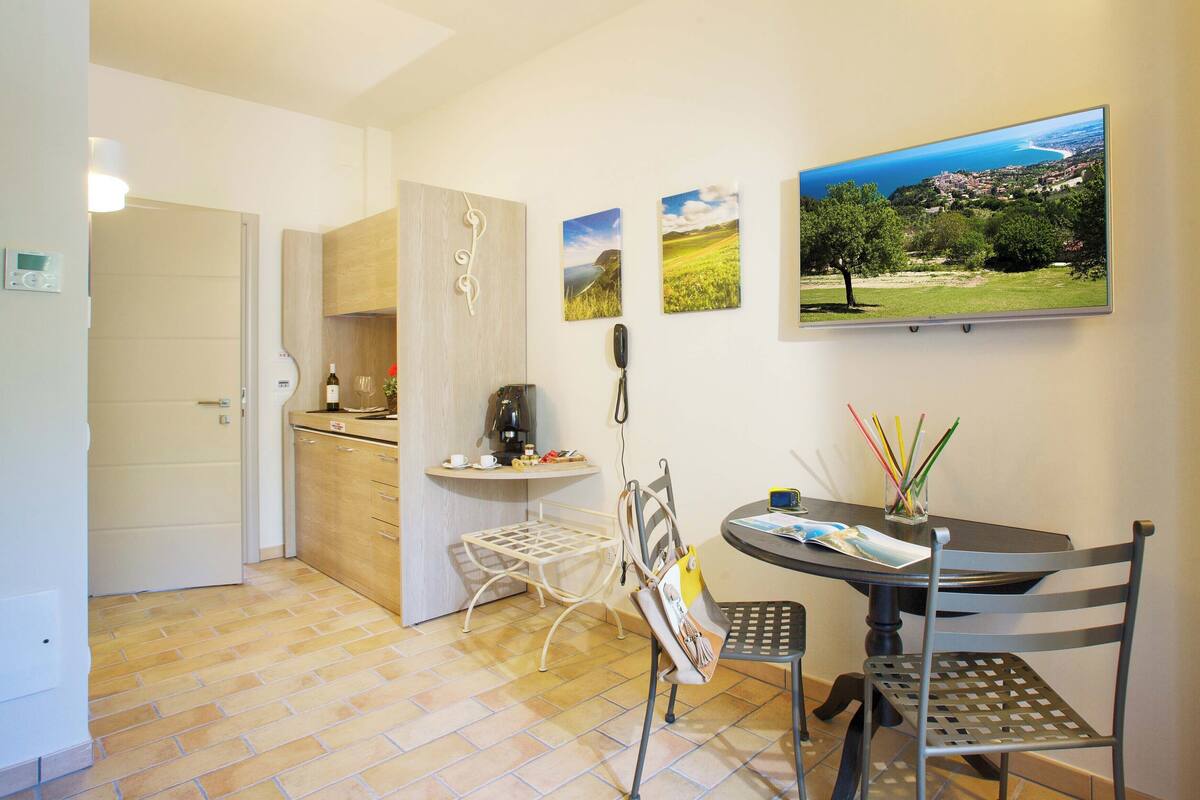 Une pièce comprenant une table à manger, des chaises, un petit coin cuisine et un téléviseur diffusant un paysage.