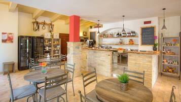 A kitchen with a central island, wooden tables, and chairs. There are refrigerators, shelves with bottles, and a chalkboard sign.