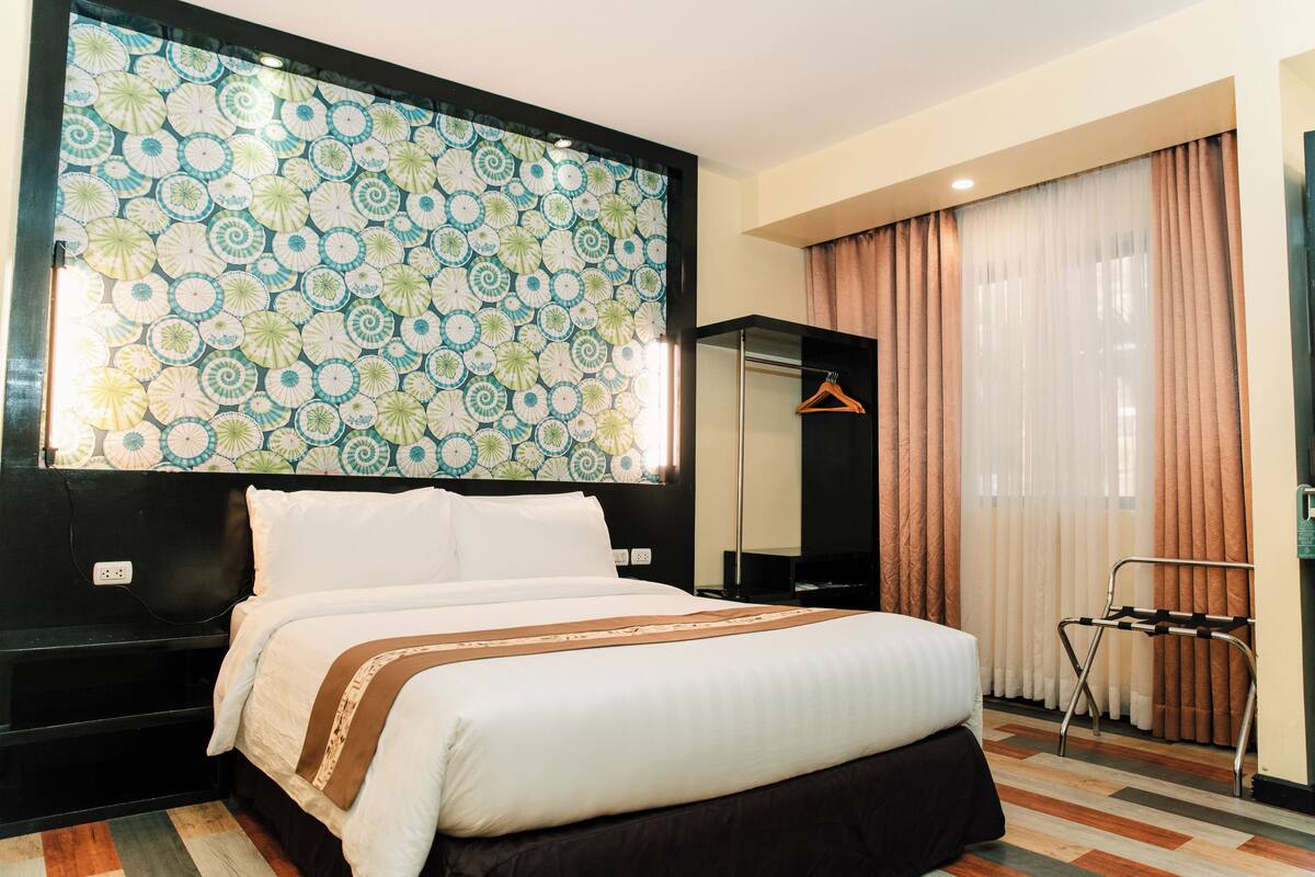 A hotel room with a large bed, a headboard with a patterned wallpaper, a mirror, a hanging coat rack, and a window with curtains.