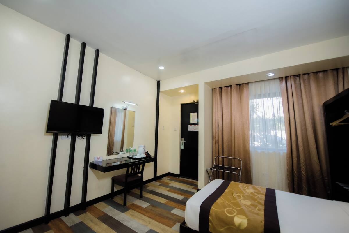 A hotel room with a bed, a desk with a chair, a flat-screen TV mounted on the wall, and a window with curtains.
