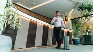 A man standing at a reception desk in a modern hotel lobby with a large potted plant and a clock on the wall. - Sun City Suites (General Santos)