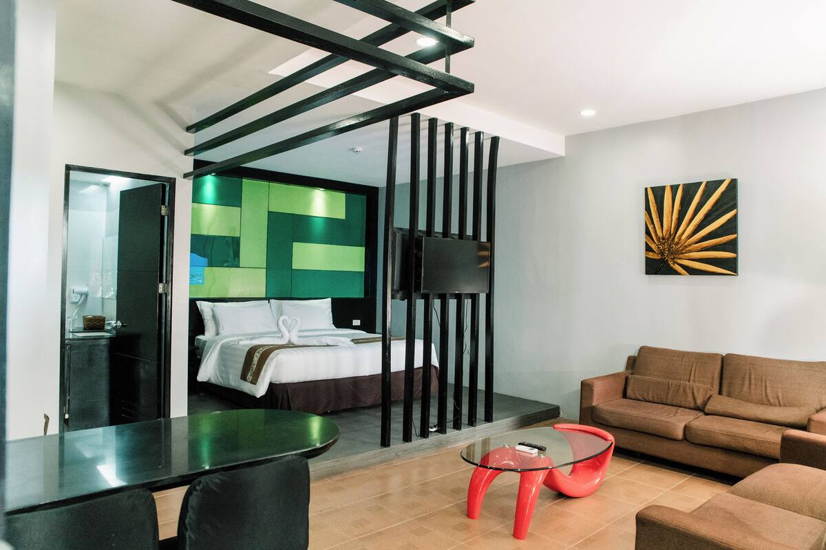 A modern hotel room with a bed, a sofa, a glass coffee table, and a TV mounted on the wall.