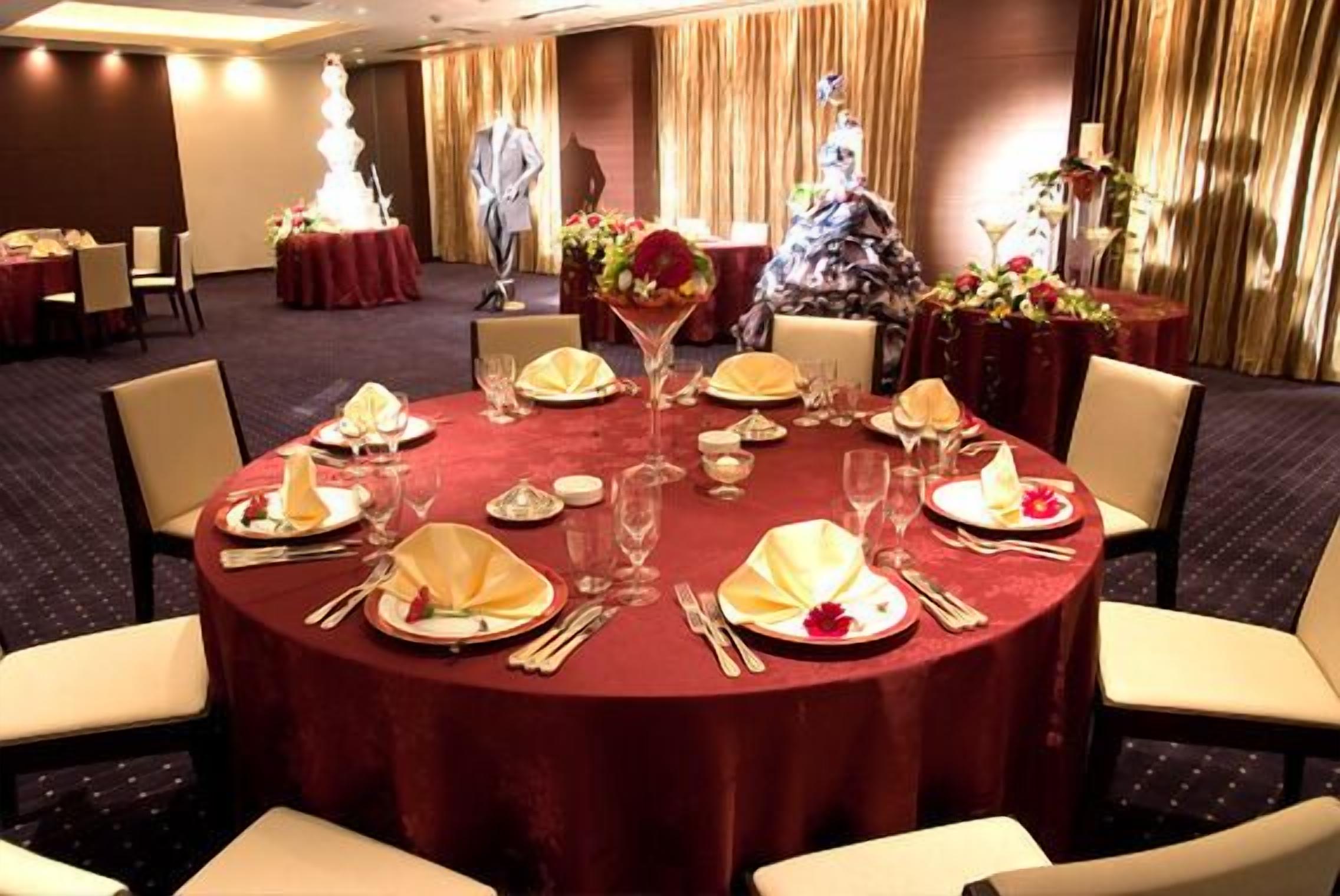 Mesa redonda posta para um jantar formal, com toalha de mesa vermelha, guardanapos brancos e talheres dourados.