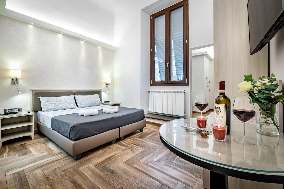 Une chambre moderne avec un lit, des tables de chevet, une table en verre avec des verres à vin et une bouteille, et une grande fenêtre avec des stores.