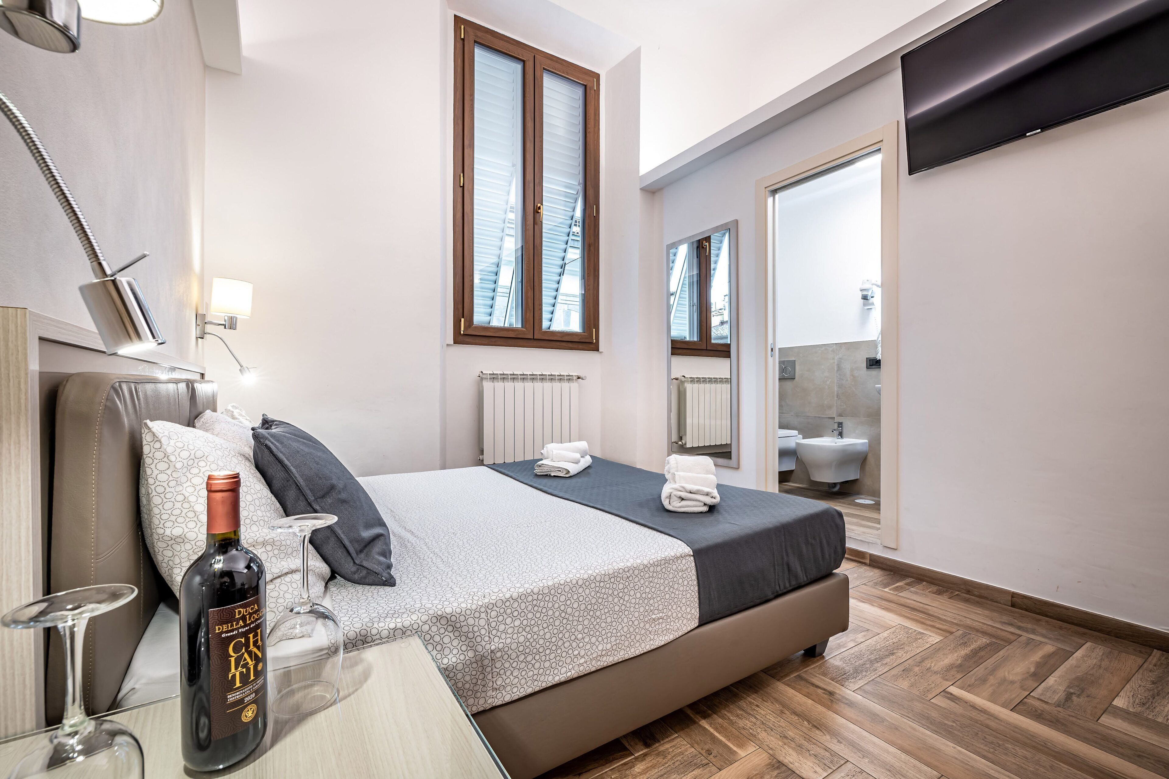 Una habitación de hotel moderna con una cama, una mesita de noche con una botella y vasos, una ventana con persianas, un espejo y un baño visible a través de una puerta abierta.
